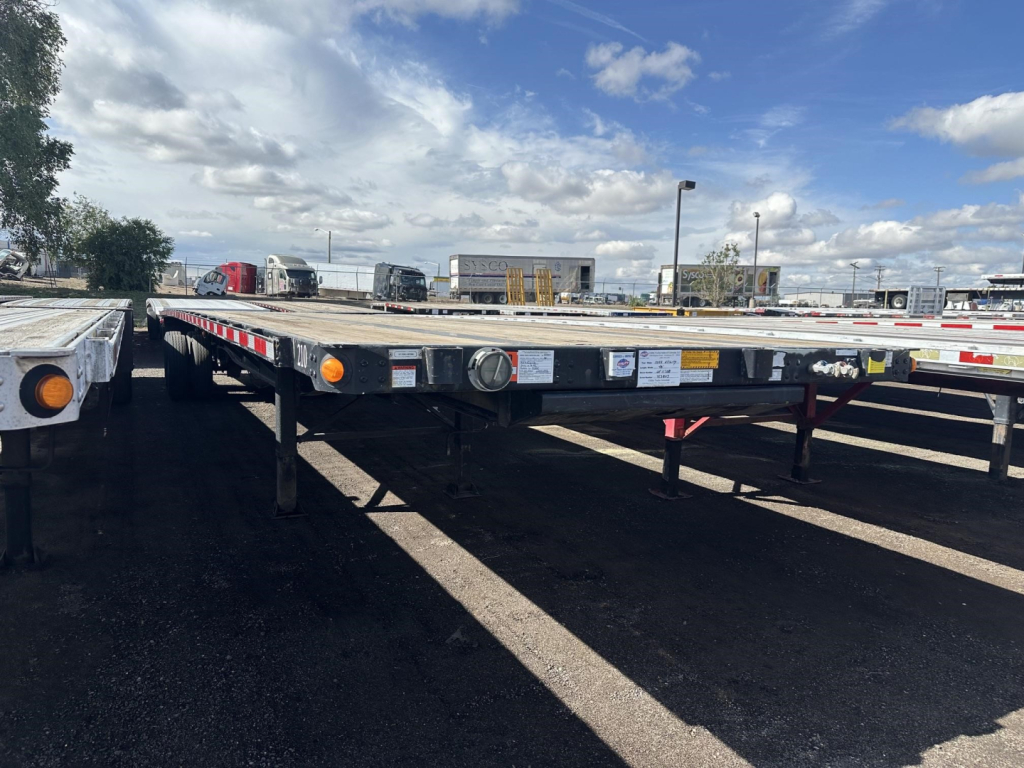 Flatbed trailer showing front end with warning labels and reflectors along the side, under a blue sky with clouds.