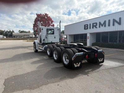 Semi truck chassis with triple rear axles and fifth-wheel, white cab, parked in lot in front of a building with BIRMIN... signage visible; no visible license plates or serials.