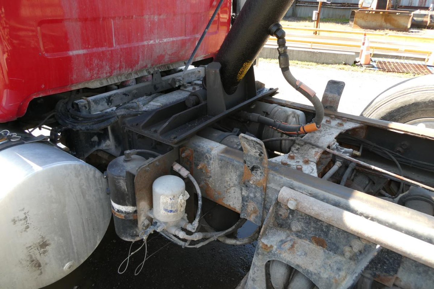 Truck chassis area showing hydraulic lines and white storage/filters mounted near the frame with a red cab above; metal framework and cylindrical aluminum component visible.