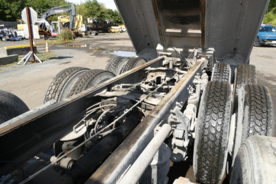 Truck chassis and dump bed area showing dual rear axles and suspension components with hydraulic rams and undercarriage. No legible brand text visible from this angle.