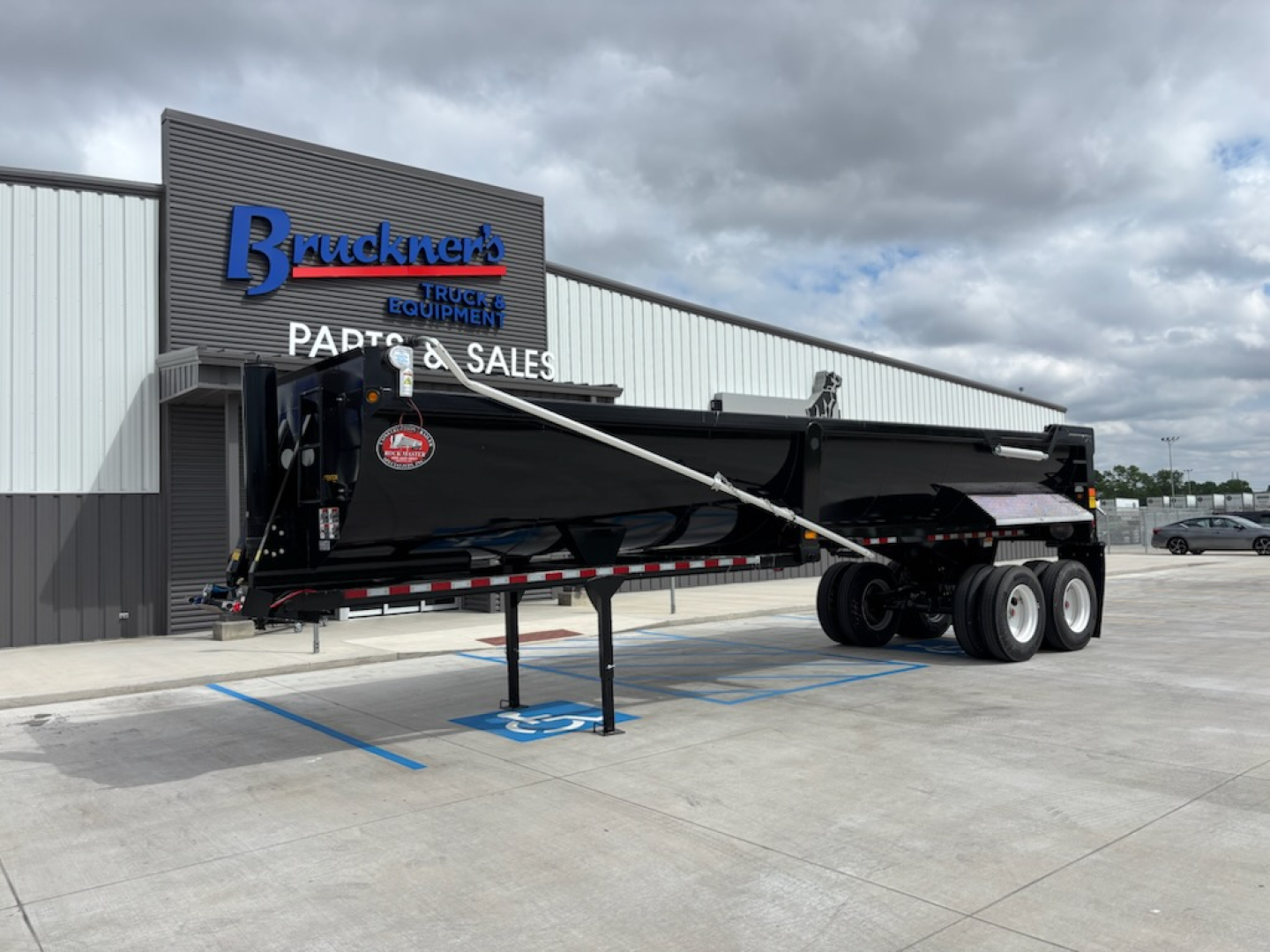 Dump trailer in a dealership lot showing a black gooseneck trailer with tandem axles and landing gear, parked in front of a building with Buckner's Trucks & Equipment sign.
