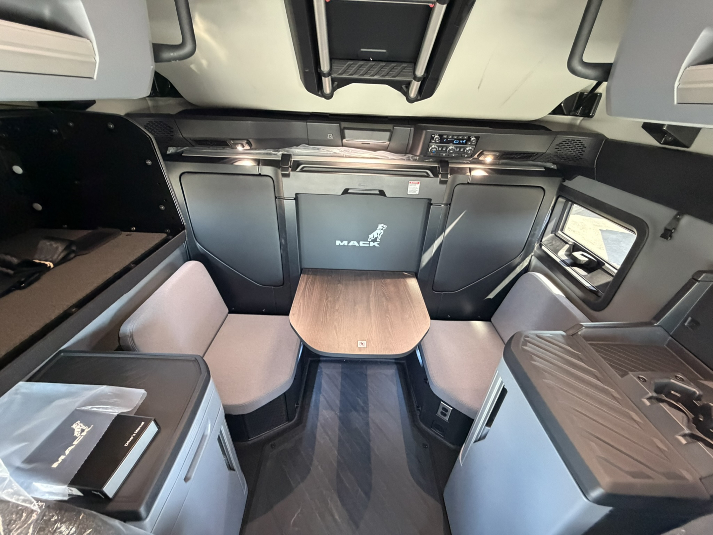 Interior of a new Mack PR64T76H sleeper cab showing front seating area and table, with Mack logo on center cabinet.