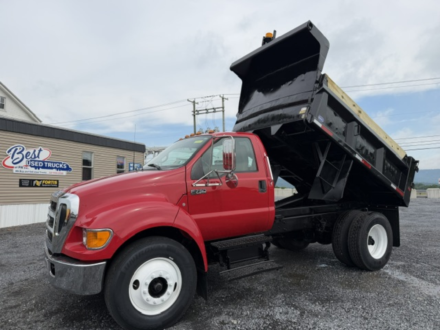 Dump truck with red cab and raised black dump bed, Ford badge visible, appears to be a 2006 Ford F-750 dump truck in a used truck lot.