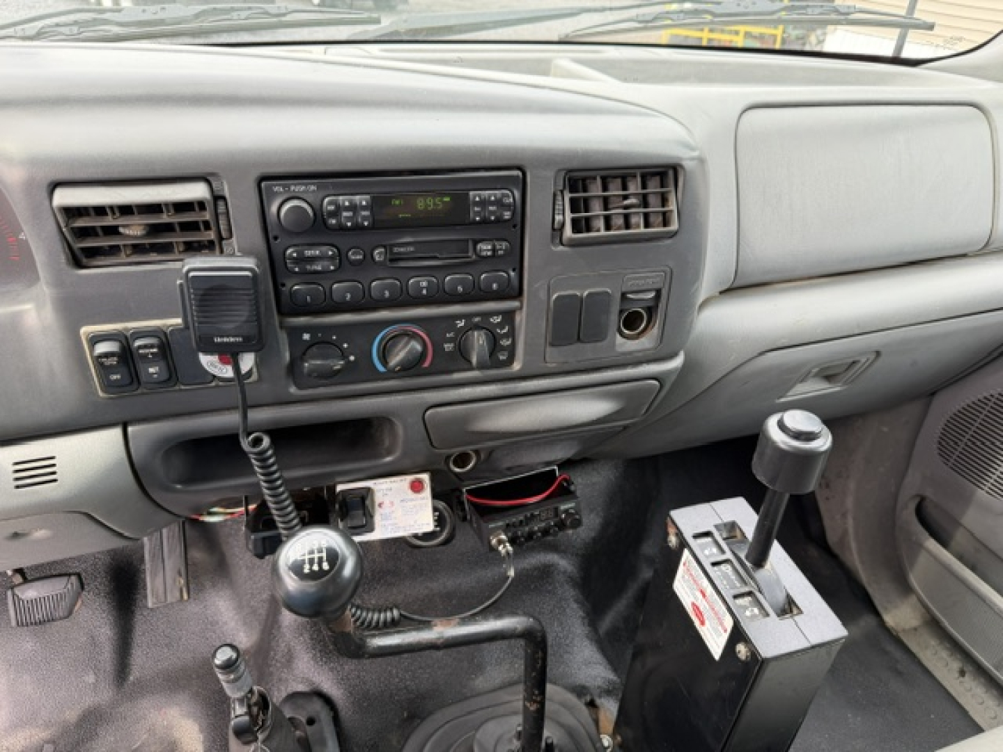 Ford F-750 interior dashboard and gear shifter area with radio, climate controls, and a mounted device; manual transmission with gear stick and another lever in front; auxiliary battery and wiring visible under dash.