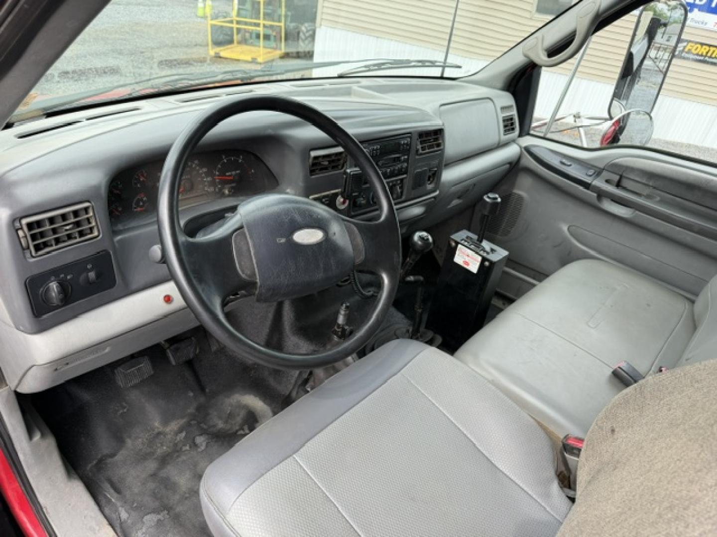 Ford F-750 interior cab view showing steering wheel, dashboard, and driver seat; grey fabric seats, manual gear shift, and worn floor area.