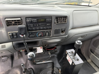 Ford F-750 interior dashboard and gear shifter area with radio, climate controls, and a mounted device; manual transmission with gear stick and another lever in front; auxiliary battery and wiring visible under dash.