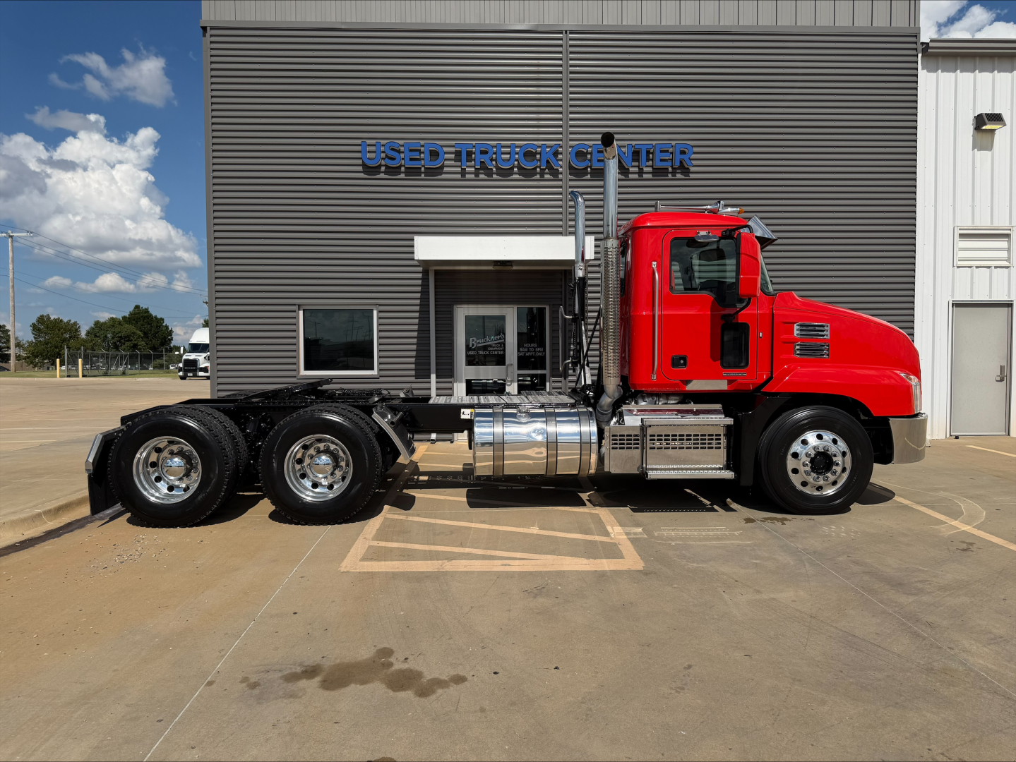 2022 Mack Anthem conventional daycab truck side view showing red cab, chrome fuel tanks, and dual rear axles in front of used truck center.