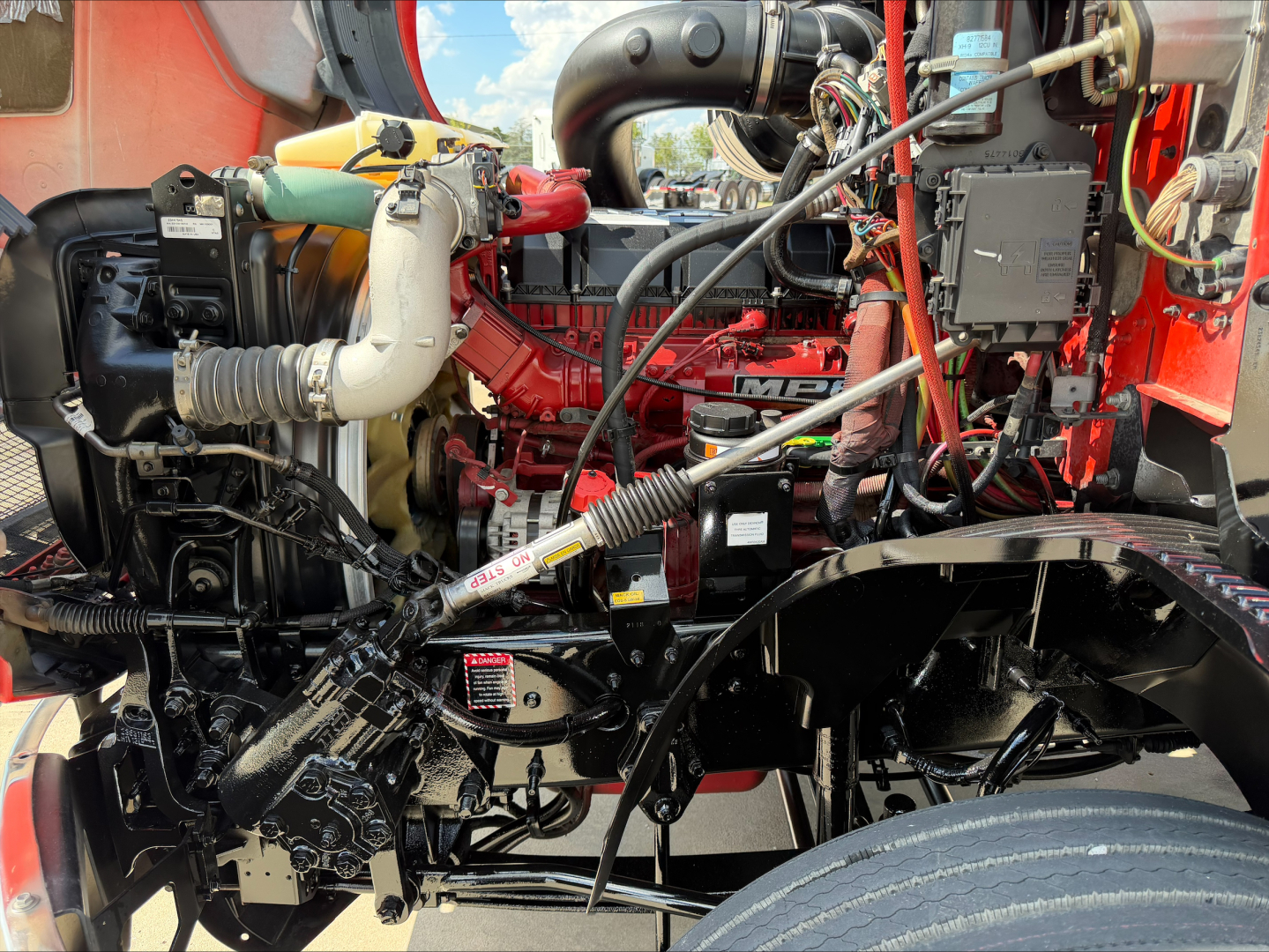Mack ANTHEM engine bay view showing red engine, hoses, wiring, and fluid reservoirs; various components and belts visible.