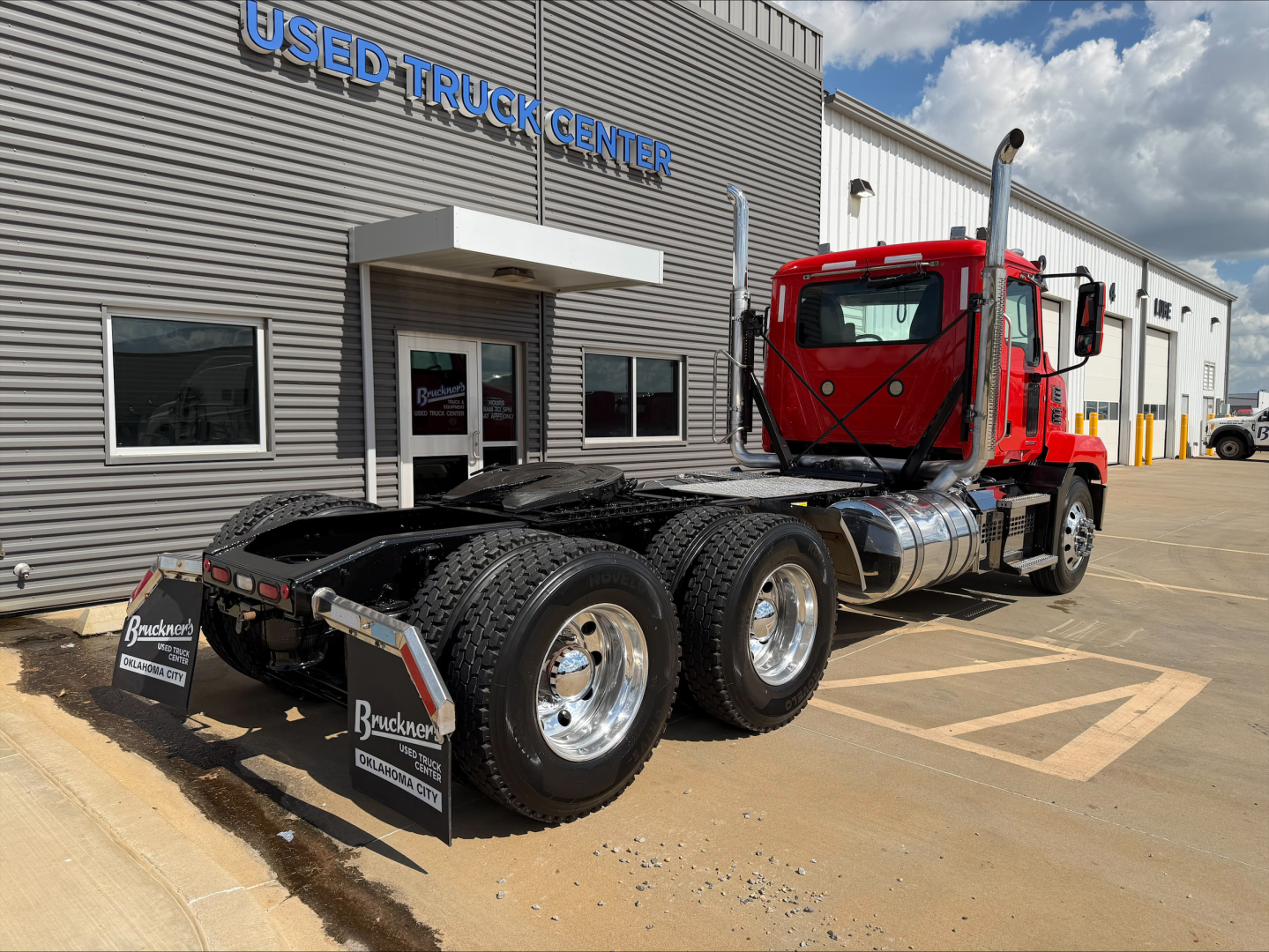 Mack Anthem tractor on lot facing camera, red cab, dual rear axles, fuel tanks, and chrome wheels beside Used Truck Center building.