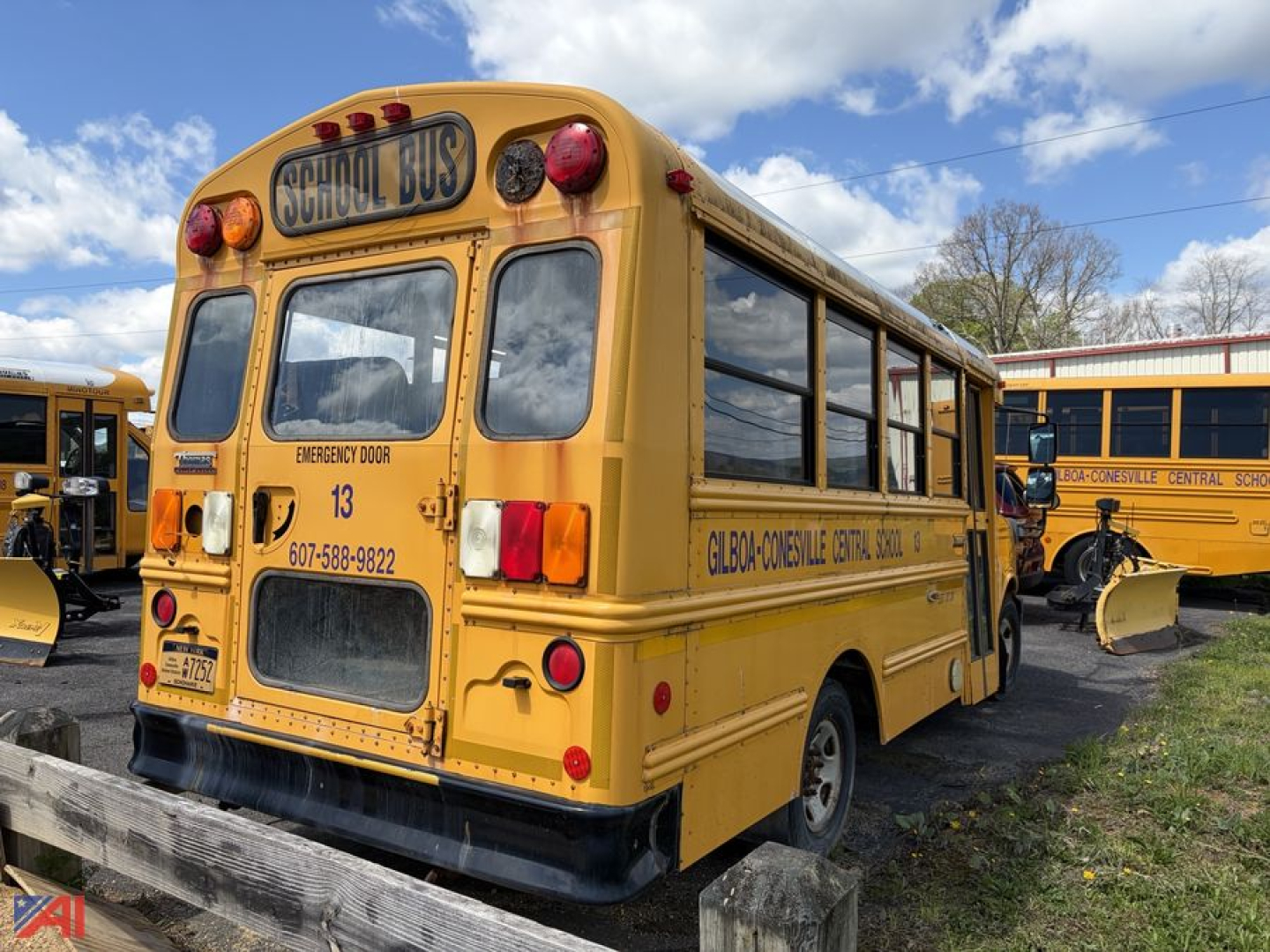 Rear view of a yellow Thomas school bus with 'SCHOOL BUS' sign, emergency exit door, and contact numbers on the side; other buses in background.