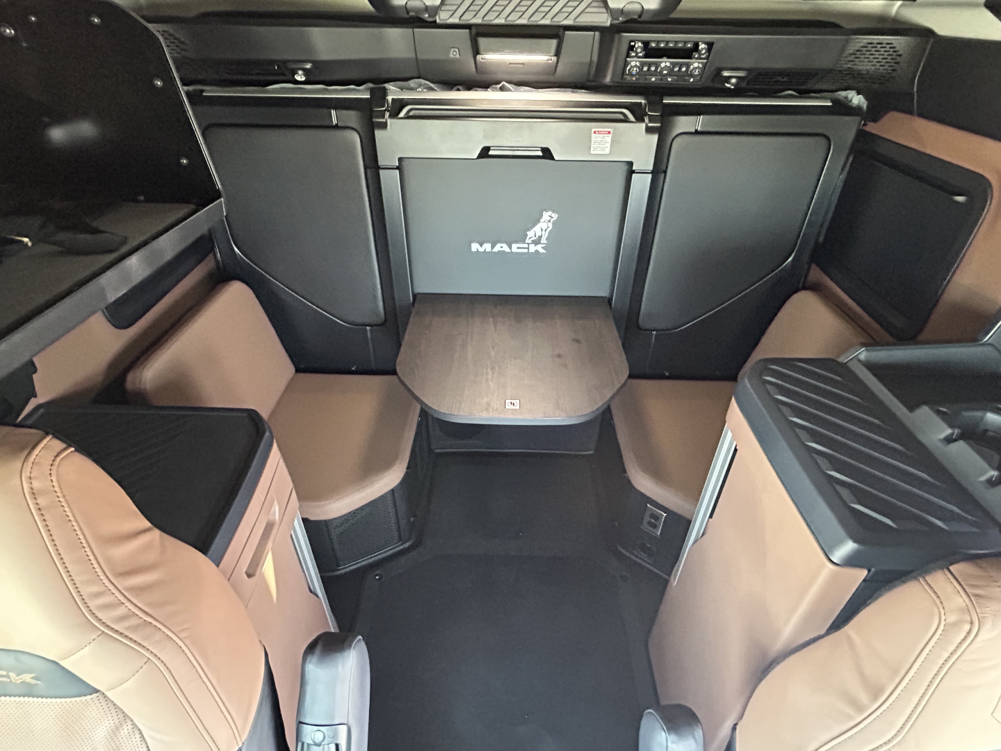 Cab interior of a 2027 Mack PR64T76H sleeper showing tan leather seats, compact table, and dashboard with storage compartments.
