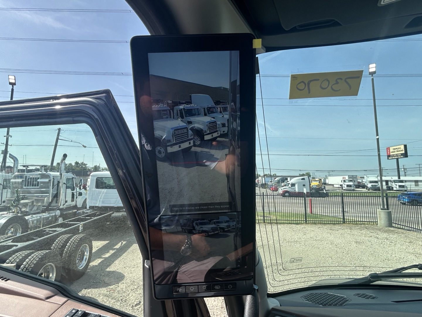 Truck cab interior showing a vertical tablet-like device mounted on the dashboard with a reflection of a truck lot in the screen window; background shows trucks and a sign reflected in the glass.