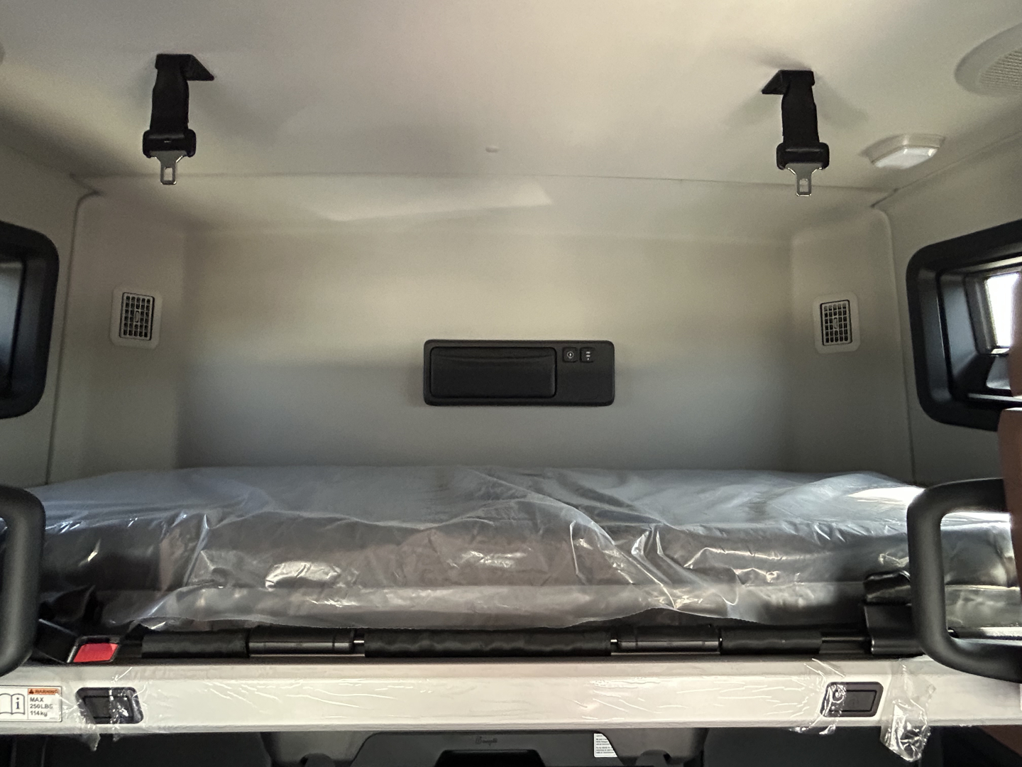 Mack PR64T76H sleeper cab interior showing bunk bed with plastic-wrapped mattress and safety belts.