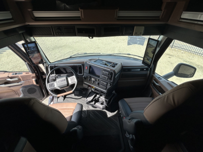 Truck cab interior with large dashboard, steering wheel, and dual display screens; brown and black seating; driver and passenger seats visible; side windows and mirrors.