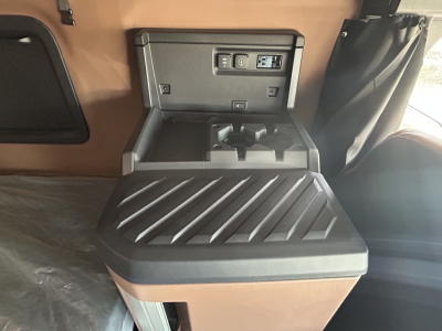 Interior view of a 2027 Mack PR64T76H cab area showing a center storage console with cup holders and control panel, brown upholstery, and a ribbed top surface.