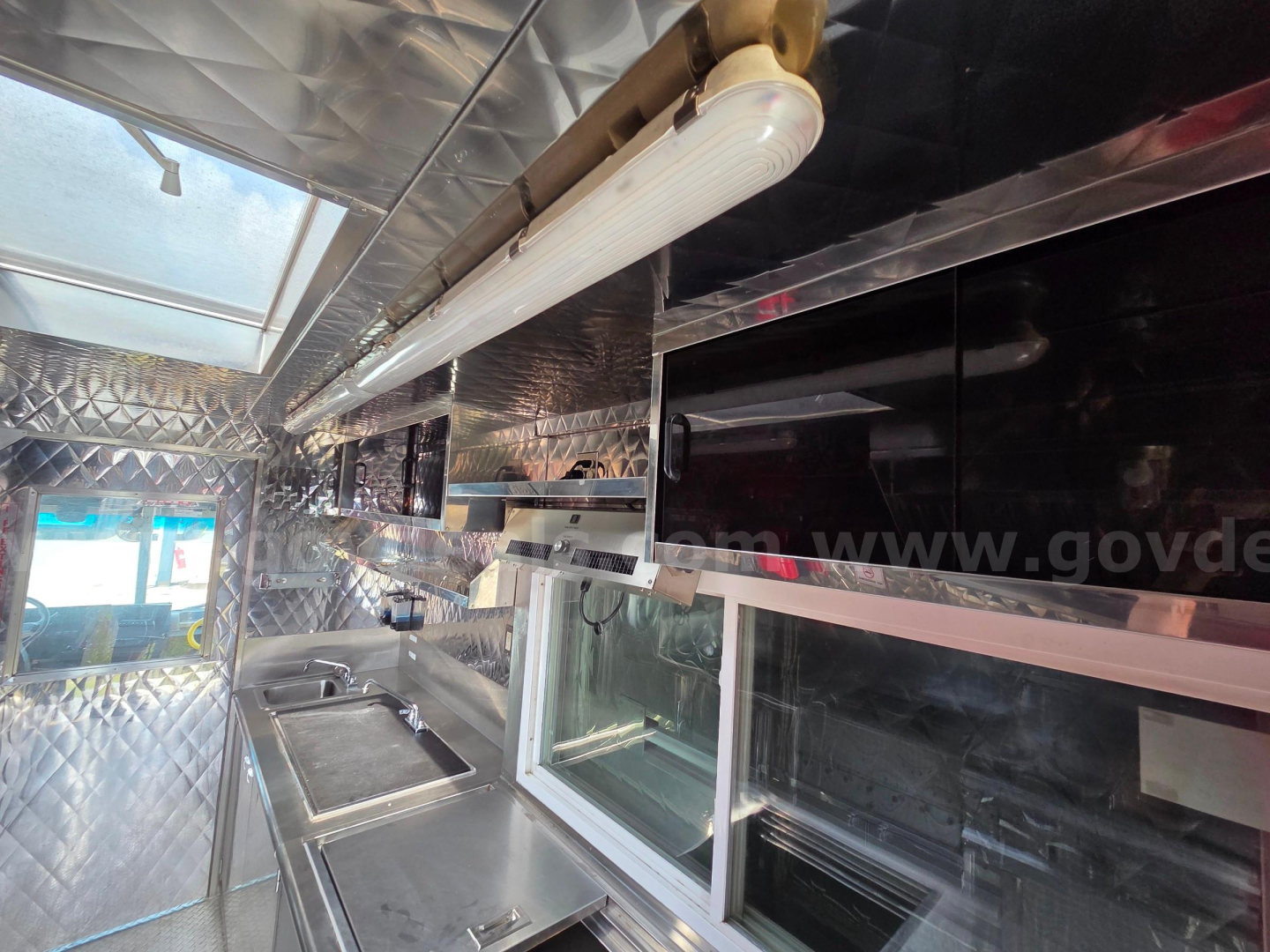 Food truck interior with stainless steel prep counter, cabinets, and luminous lighting fixture along the ceiling; diamond plate walls and serving window visible.