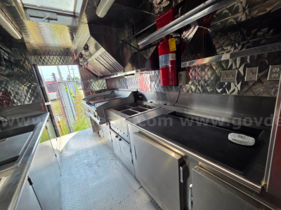 Truck interior showing stainless steel food prep area with countertop, sink, storage drawers, and a fire extinguisher mounted; aluminum diamond plate walls and ceiling; kitchen equipment along both sides.