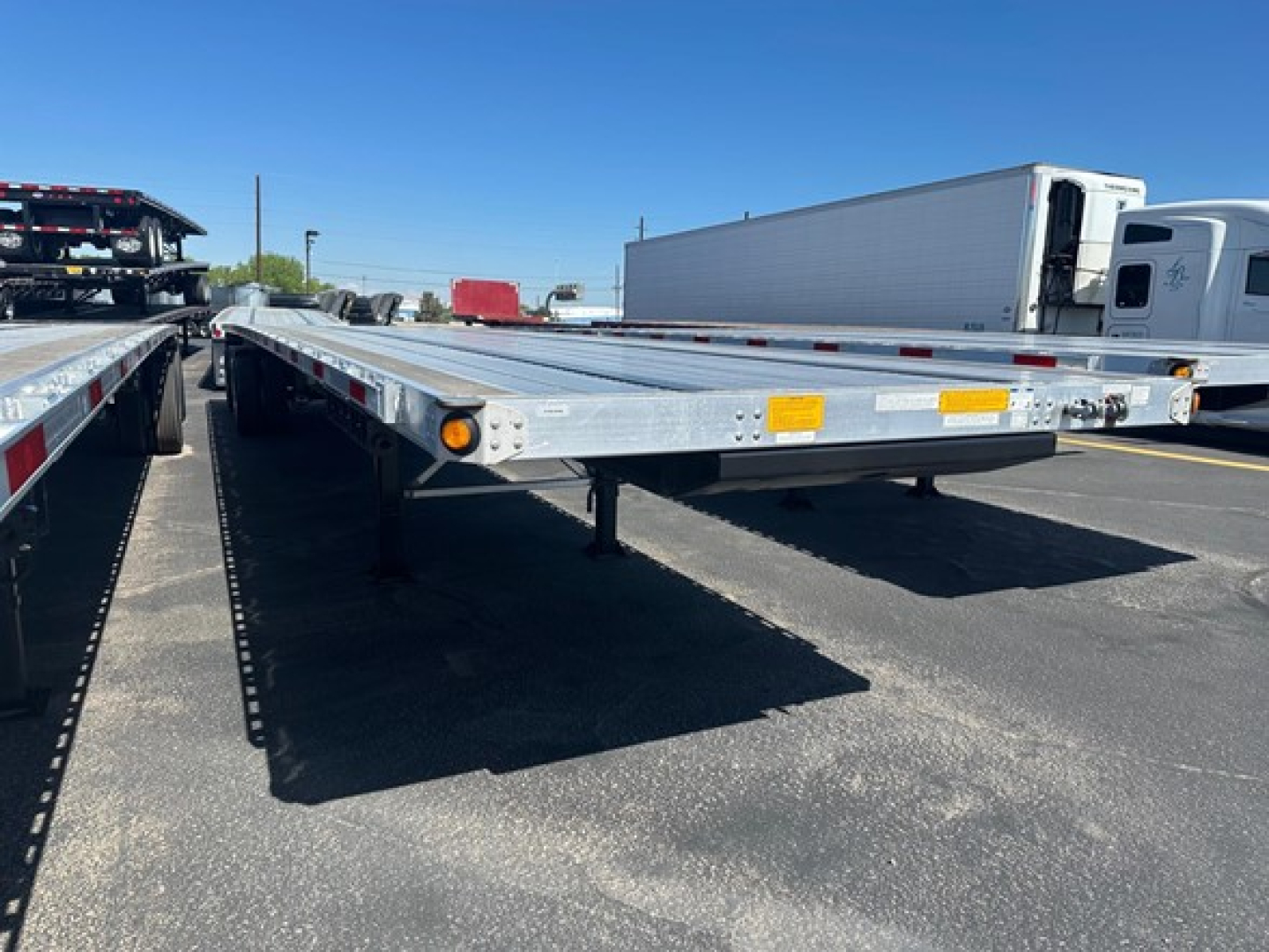Flatbed trailer with low-profile deck showing reflective markers and support legs, long metal frame, over a paved lot.