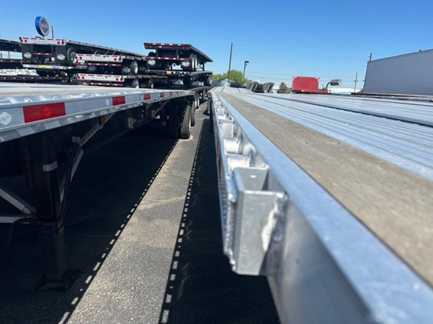 Flatbed trailer edge and deck showing aluminum side rails and deck planks with a long wheelbase trailer in background.