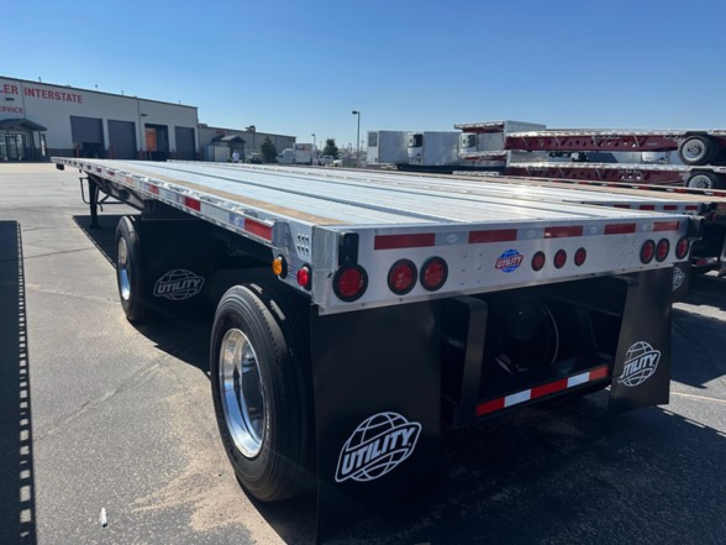 Flatbed trailer showing rear with dual axles and mudflaps marked Utility, aluminum deck, rear lighting, and logo stickers.