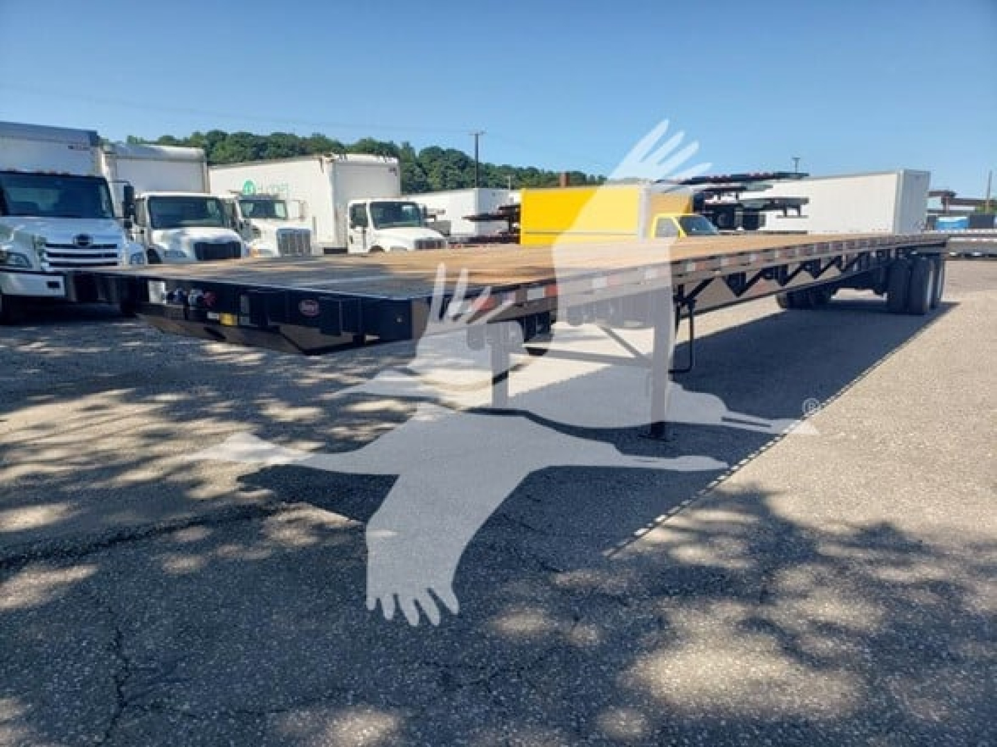 Flatbed trailer in a dealership lot showing a long black frame with wooden deck and dual rear axles.