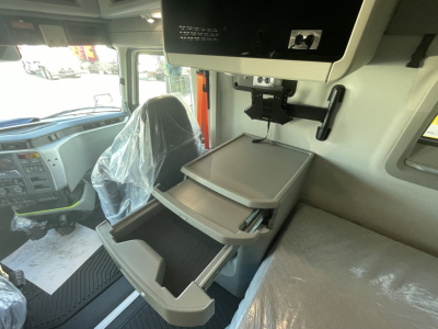  Volvo VNL64T860 cabin area showing a overhead screen and a gray fold-out storage/desk with multiple trays, driver seat with plastic cover, dashboard visible outside windows.