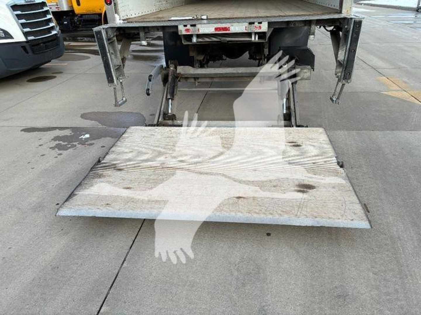 Truck rear with loading ramp deployed; metal ramp platform shows surface marks. Part of chassis and hydraulic mechanism visible beneath and behind.