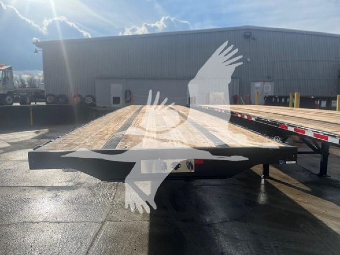 Flatbed trailer in a parking lot showing wooden deck and steel frame, with a warehouse in the background.