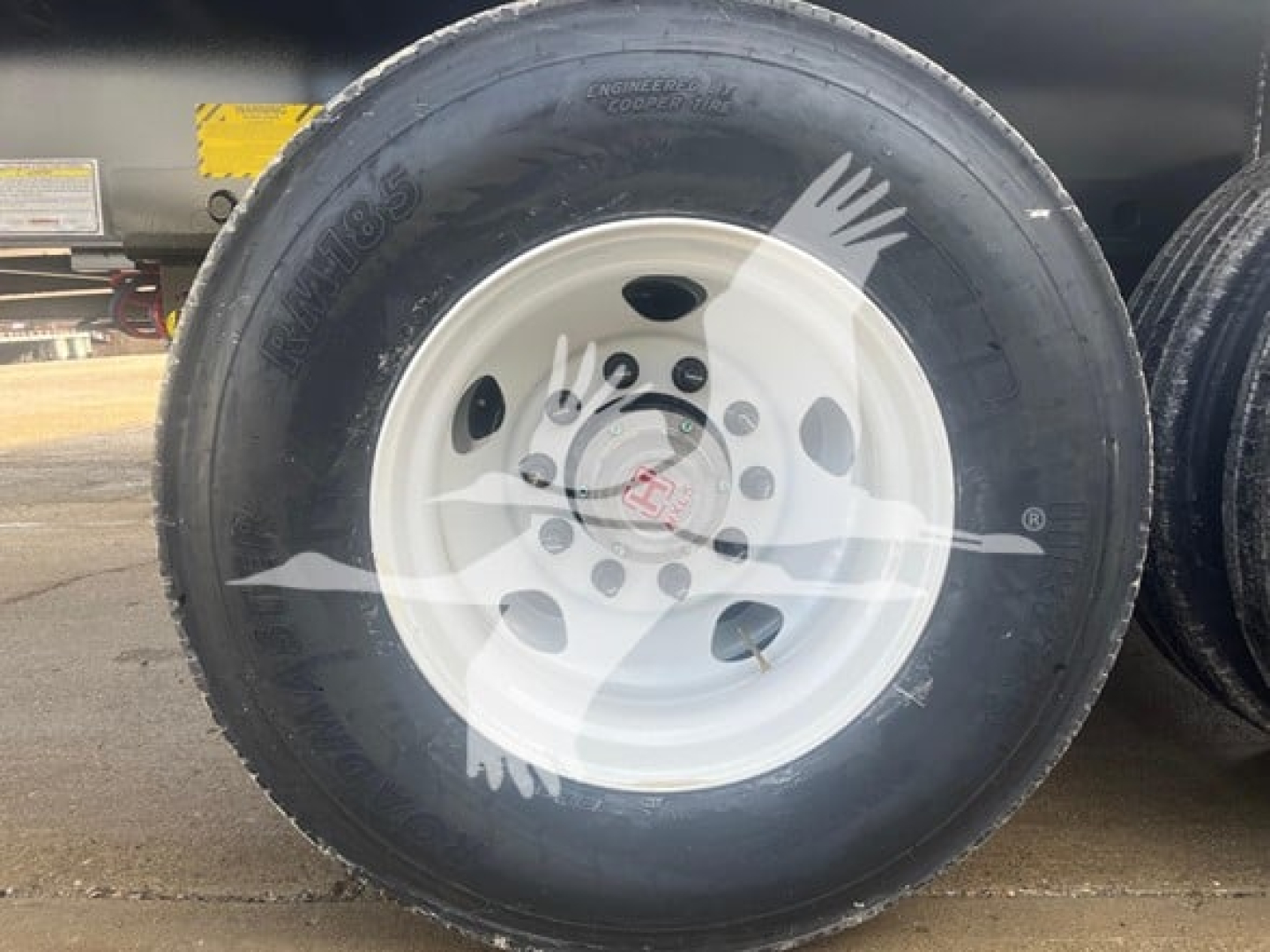 Wabash flatbed trailer wheel and tire close-up showing white steel wheel and Goodyear tire with visible tread; part of trailer undercarriage in background.
