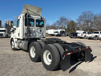 Freightliner Cascadia126 tractor rear view showing fifth wheel, dual rear axles, and mud flaps with wake behind cab; street and other trucks in background.