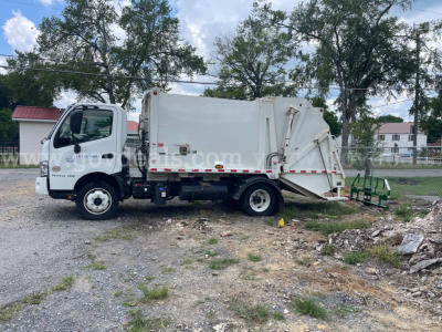 Garbage truck side view 2019 Hino 195 with rear compactor, white body, cab, and hydraulic lifting mechanism visible.