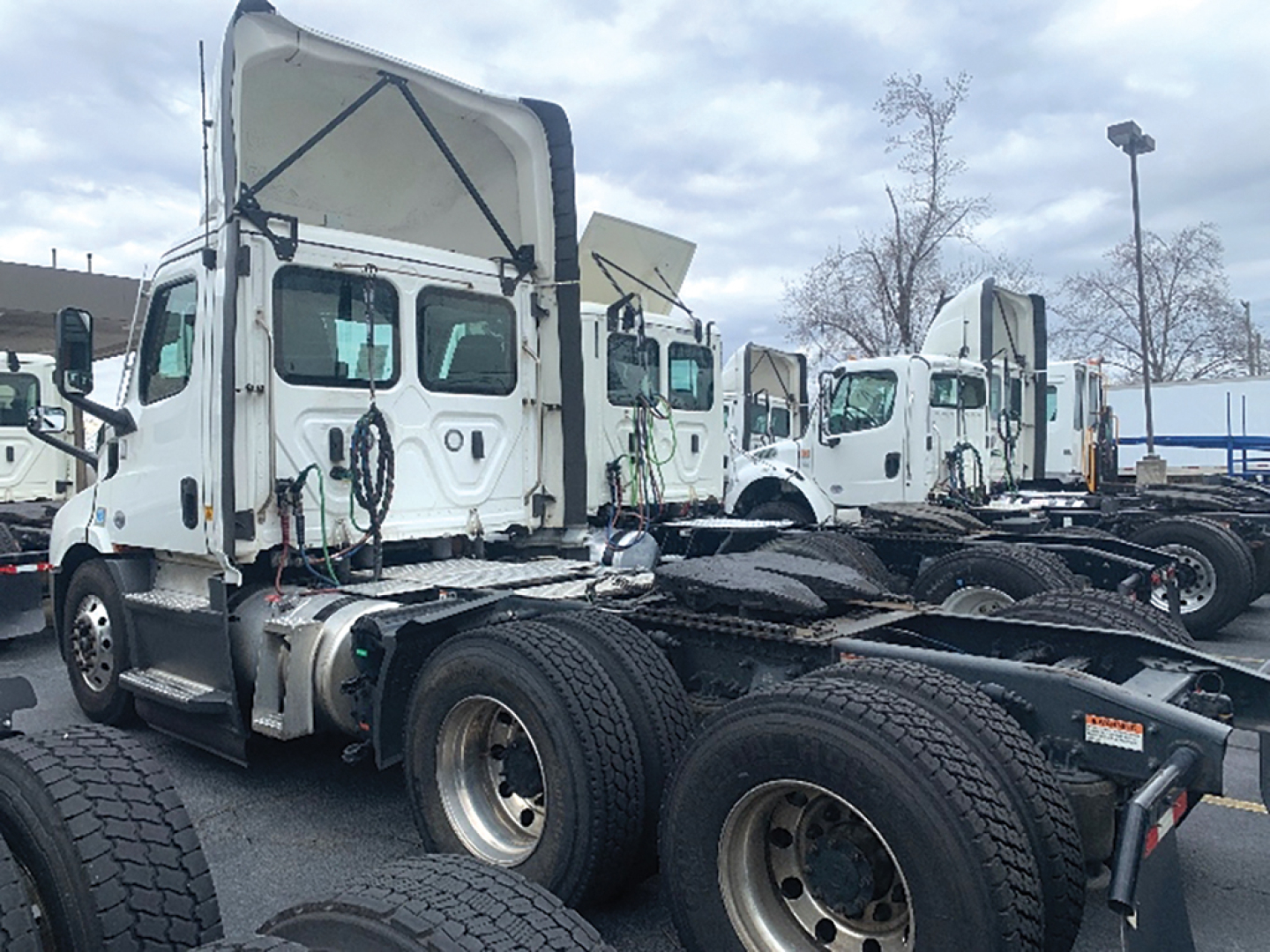 Truck tractor cab and chassis in a parking lot, white Freightliner Cascadia Daycab with raised hood and dual rear axles.