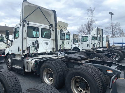 Truck tractor cab and chassis in a parking lot, white Freightliner Cascadia Daycab with raised hood and dual rear axles.