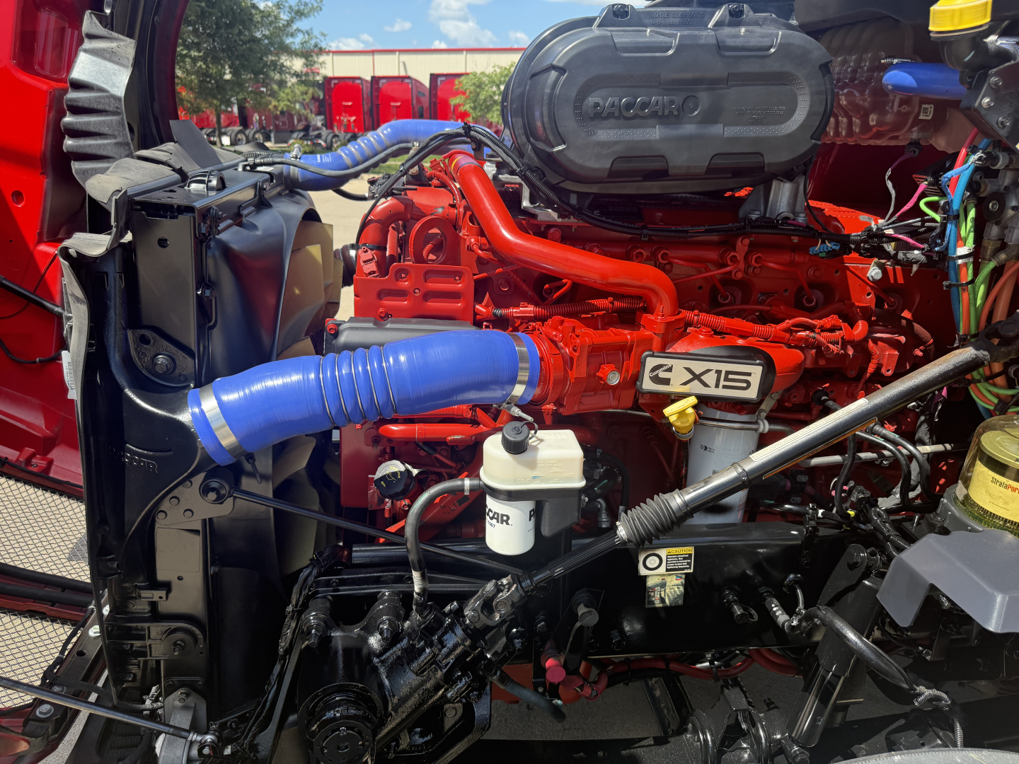 Kenworth T680 engine bay showing Cummins engine with red intake manifold, blue hoses, coolant reservoir, and various fluid reservoirs.