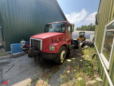 Red Freightliner FL80 cab and chassis truck parked between green metal buildings, with another yellow truck in background; visible front grille, headlights, and side mirrors.