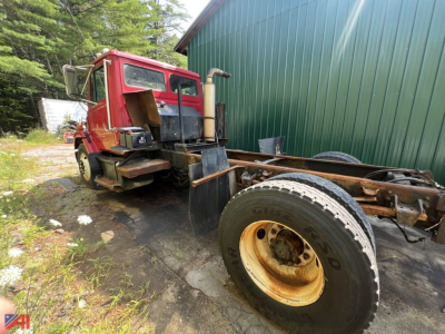 Cab and chassis truck showing Freightliner FL80 drivetrain area with fuel tank, exhaust stack, and single rear wheel, rusty frame and green building in background.