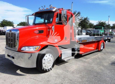 Peterbilt 337 rollback truck in red with chrome grille and flatbed; visible badge on hood, side mirrors, and steps; background shows lot with other trucks