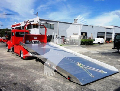 Peterbilt 337 rollback tow truck with a long aluminum ramp deployed, red cab, warehouse backdrop, clear blue sky.