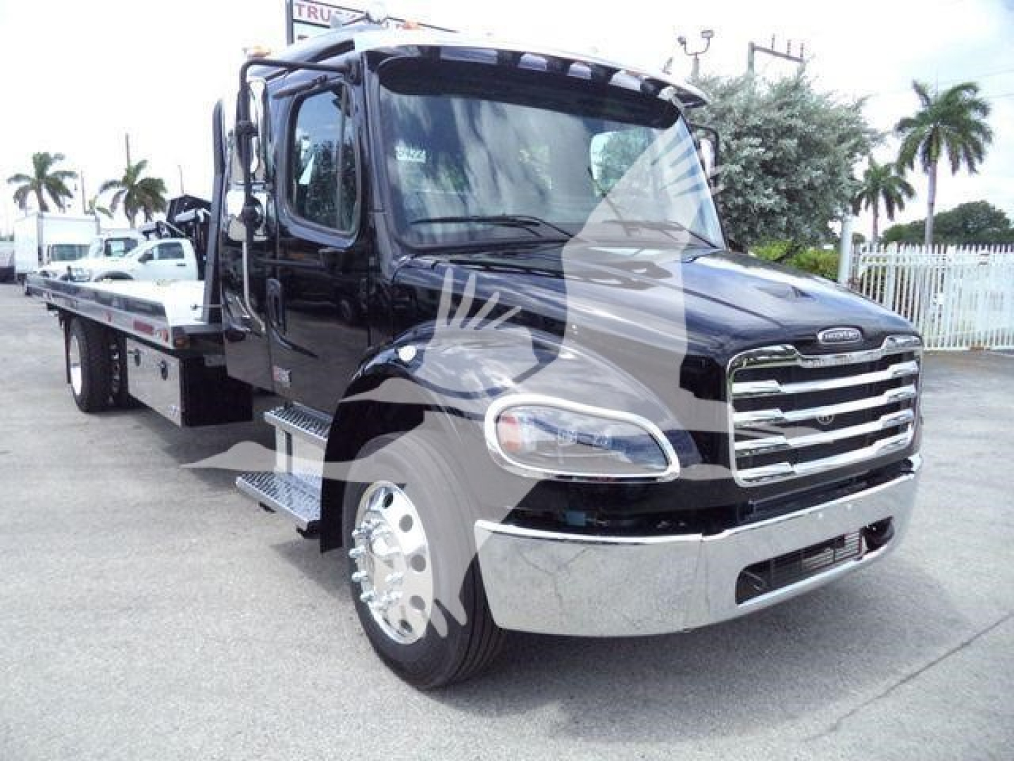 Freightliner M2 106 rollback truck in view showing cab and front grill with chrome bumper and flatbed behind.