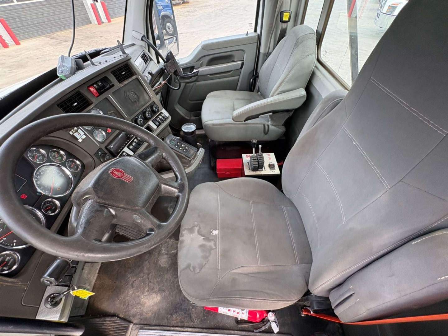 Kenworth T880 truck cabin interior showing steering wheel, dashboard gauges, driver seat with wear, center console and passenger seat; gray fabric seats; visible controls and ventilation vents.