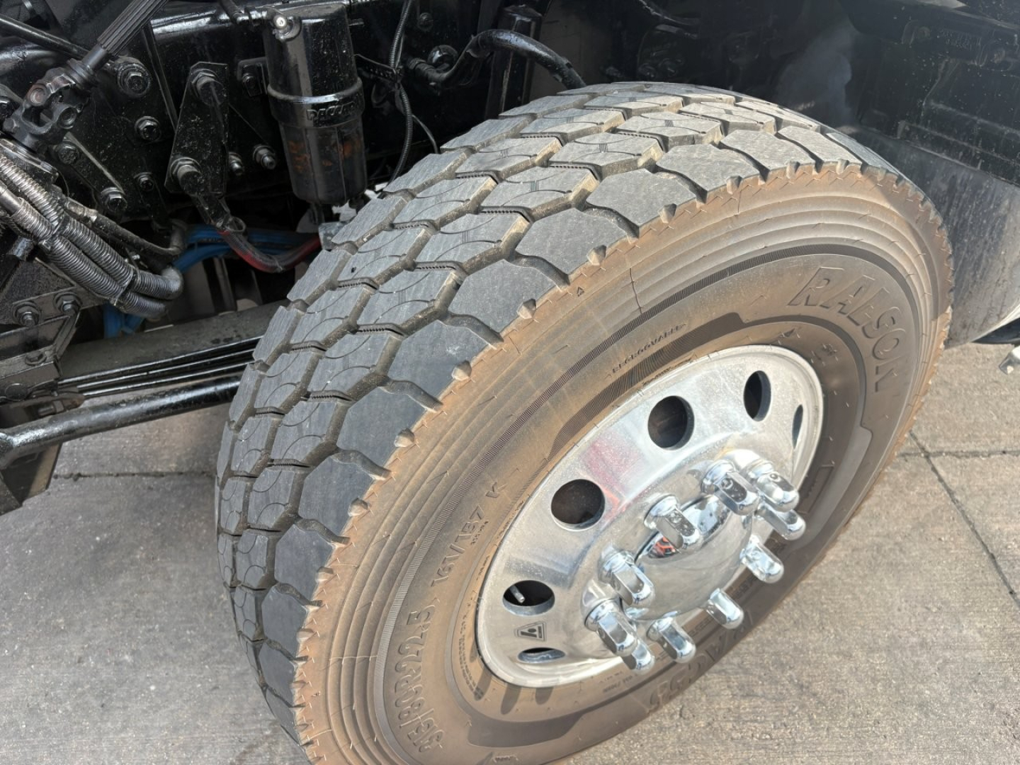 Kenworth T880 front wheel and suspension showing tire treads and chrome lug nuts, frame components and hydraulic lines nearby.