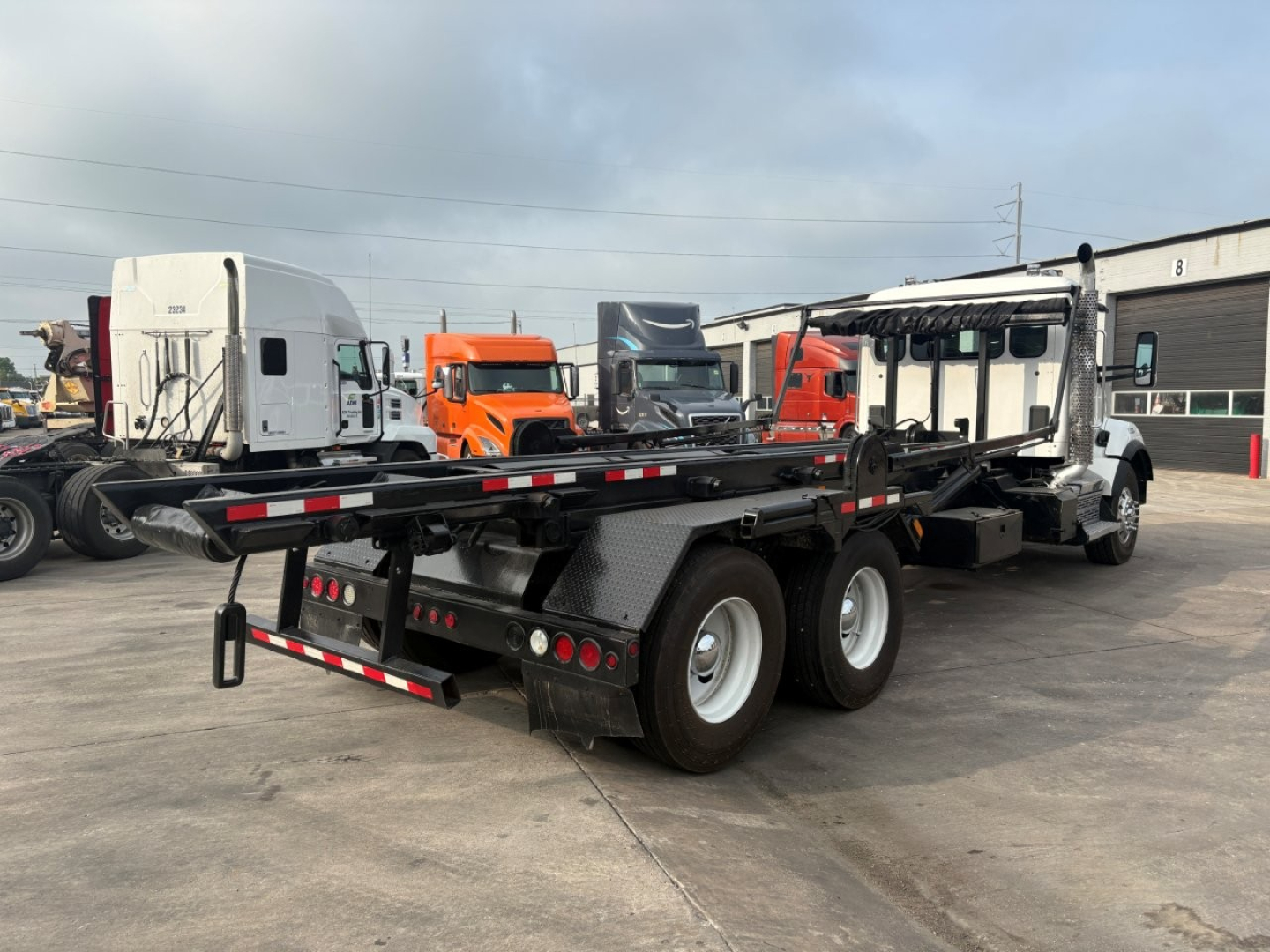 Kenworth T880 roll-off truck with flatbed and dual rear axles, viewed from rear-left side in a lot.