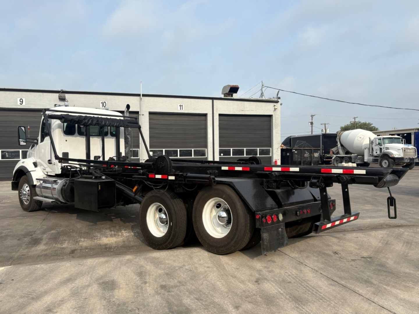 Kenworth T880 rollback truck in service with a long flatbed trailer and dual rear axles. Visible text includes stock tags and numbers on the building behind.