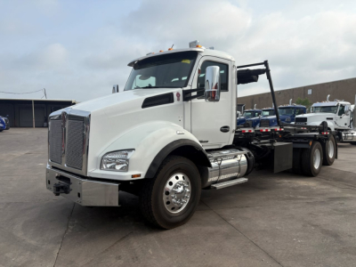 Truck image showing a white Kenworth T880 roll-off tractor with a large chrome grille, dual fuel tanks, and a flatbed rear for container hauling.