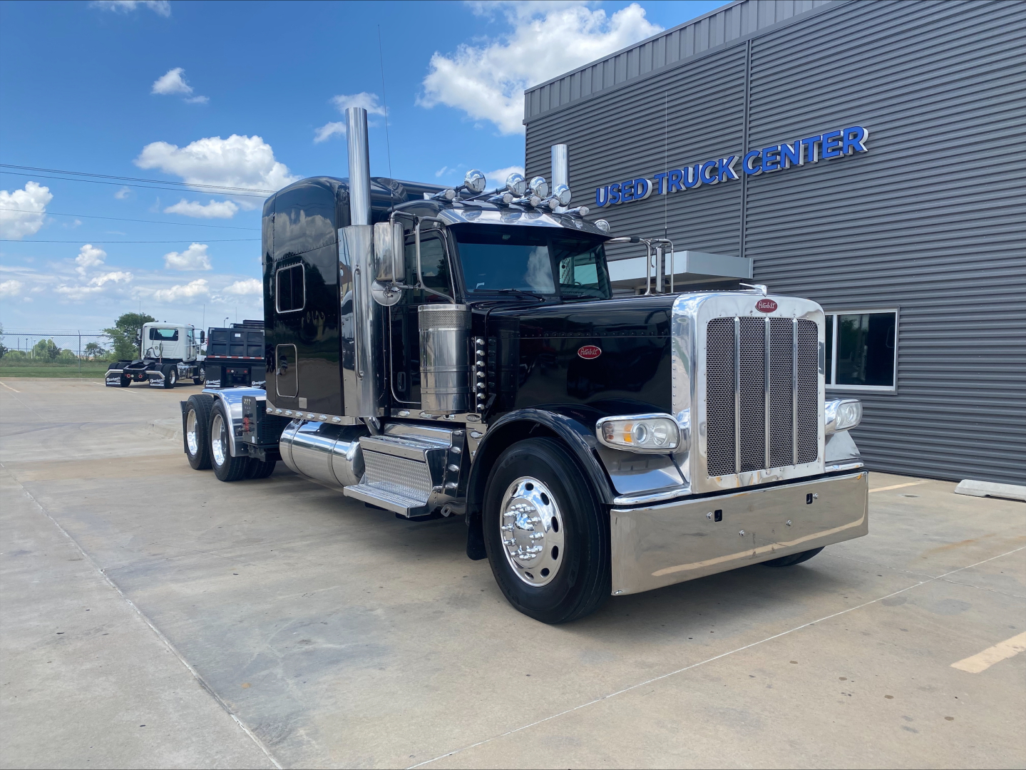 Peterbilt 389 conventional sleeper truck in front of a building, chrome bumper and exhaust stacks visible.