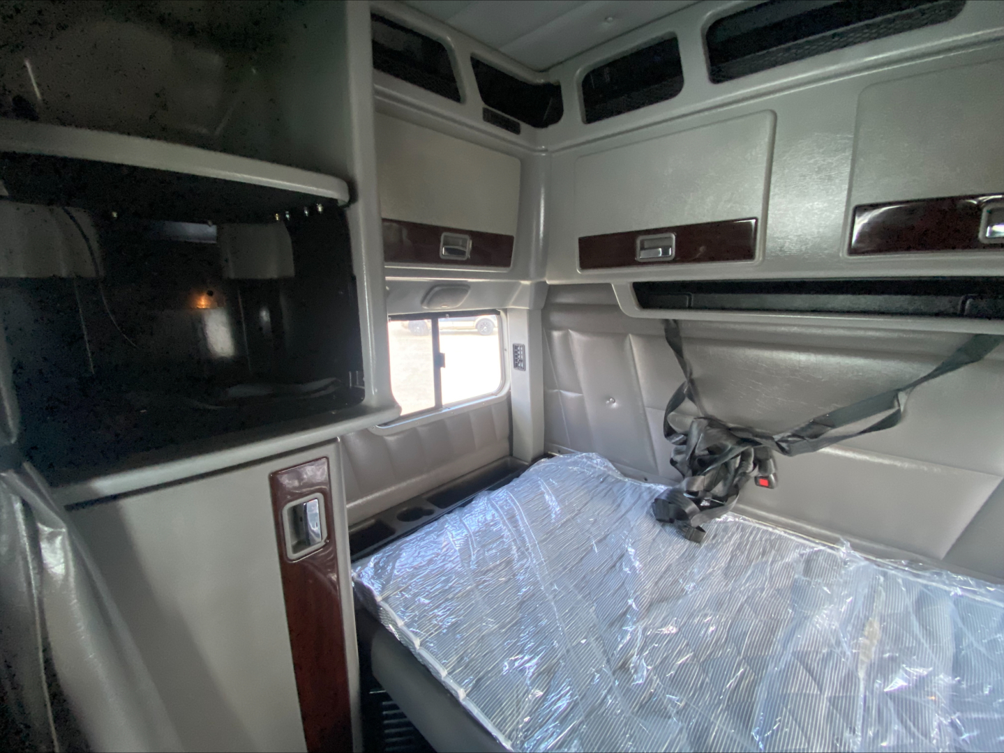 Peterbilt 389 cab interior showing sleeper bunk with plastic cover, overhead compartments, and window; beige/gray interior with wood grain trim.