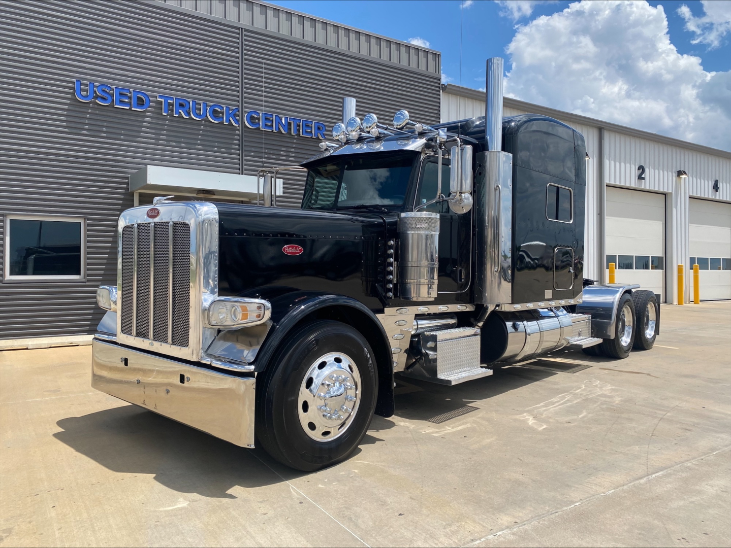 Peterbilt 389 conventional sleeper truck in a dealership lot with chrome bumper and dual exhaust stacks.