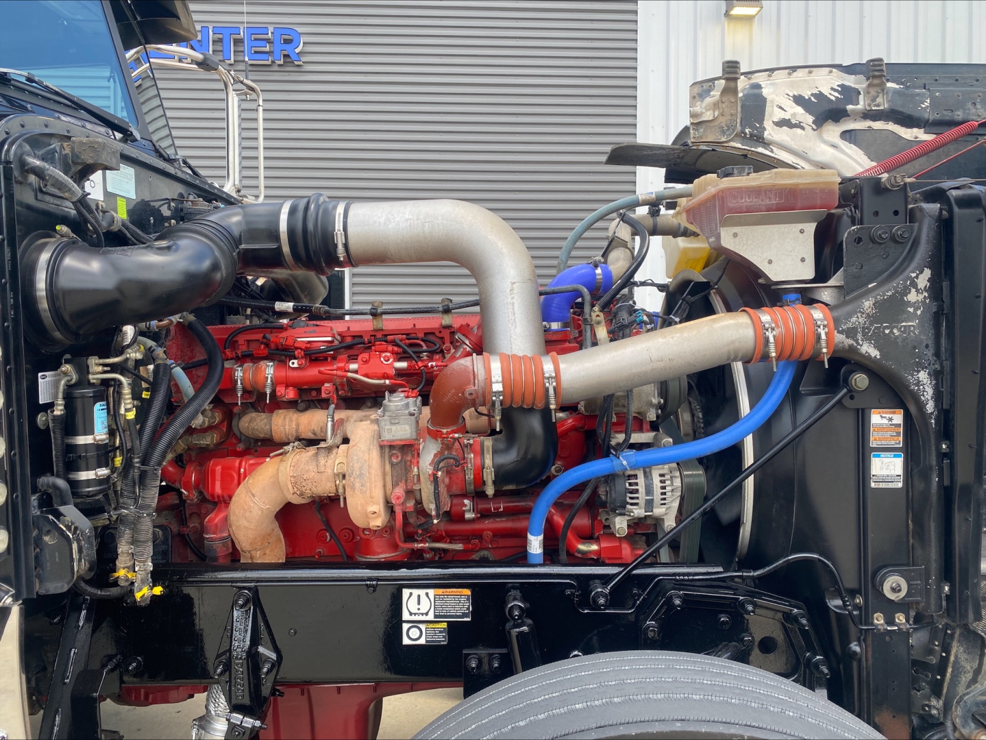 Peterbilt 389 engine bay showing red diesel engine with large aluminum intake pipe and blue hose.