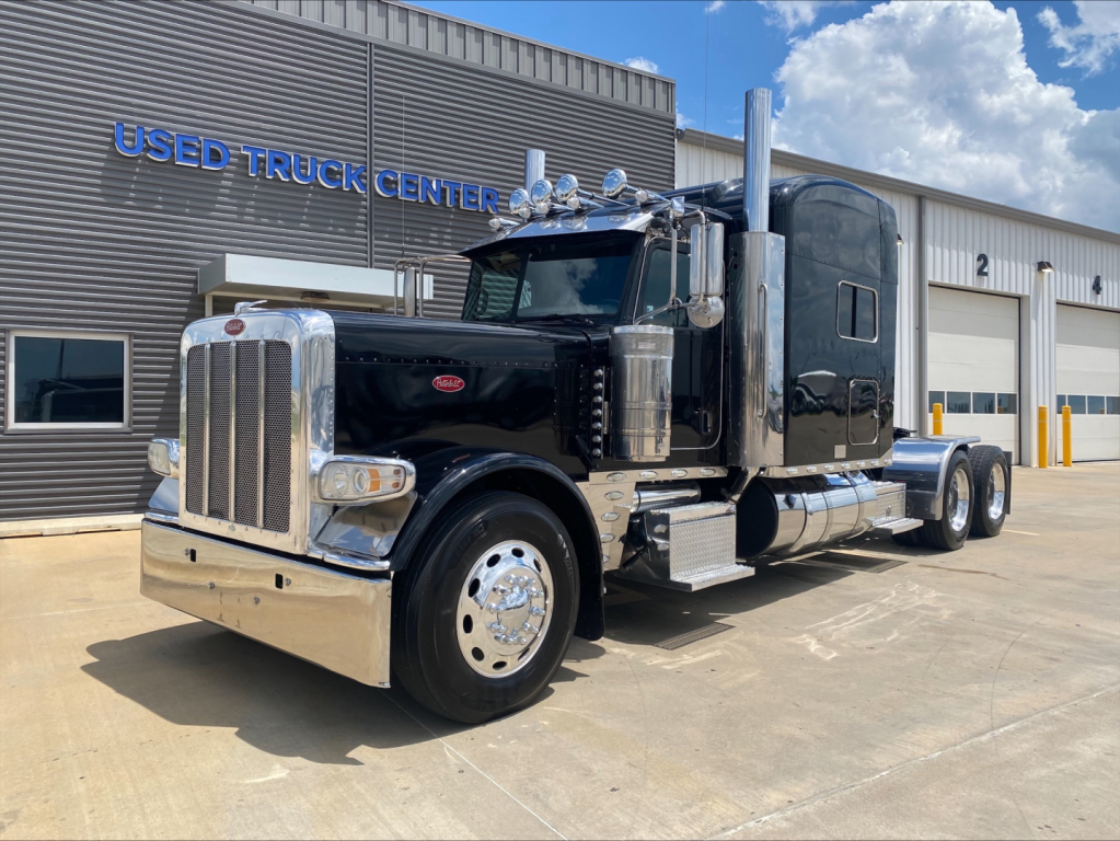 Peterbilt 389 conventional sleeper truck in a dealership lot with chrome bumper and dual exhaust stacks.