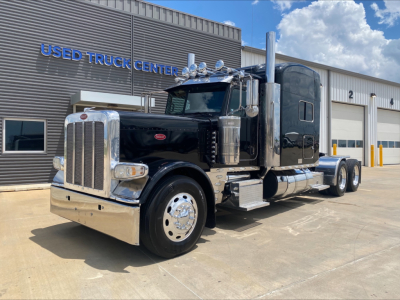 Peterbilt 389 conventional sleeper truck in a dealership lot with chrome bumper and dual exhaust stacks.